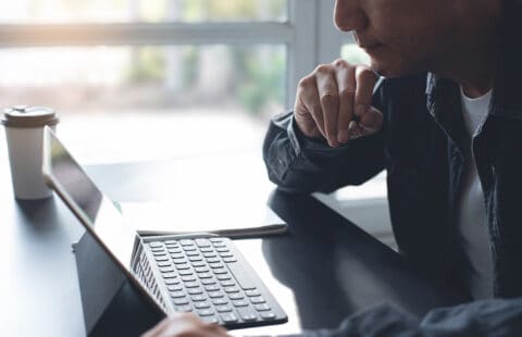 Seriously confident asian business man, freelancer in casual working and concentrated on laptop computer. Man sitting at desk in webinar via internet in modern home office with copy space, side view, close up